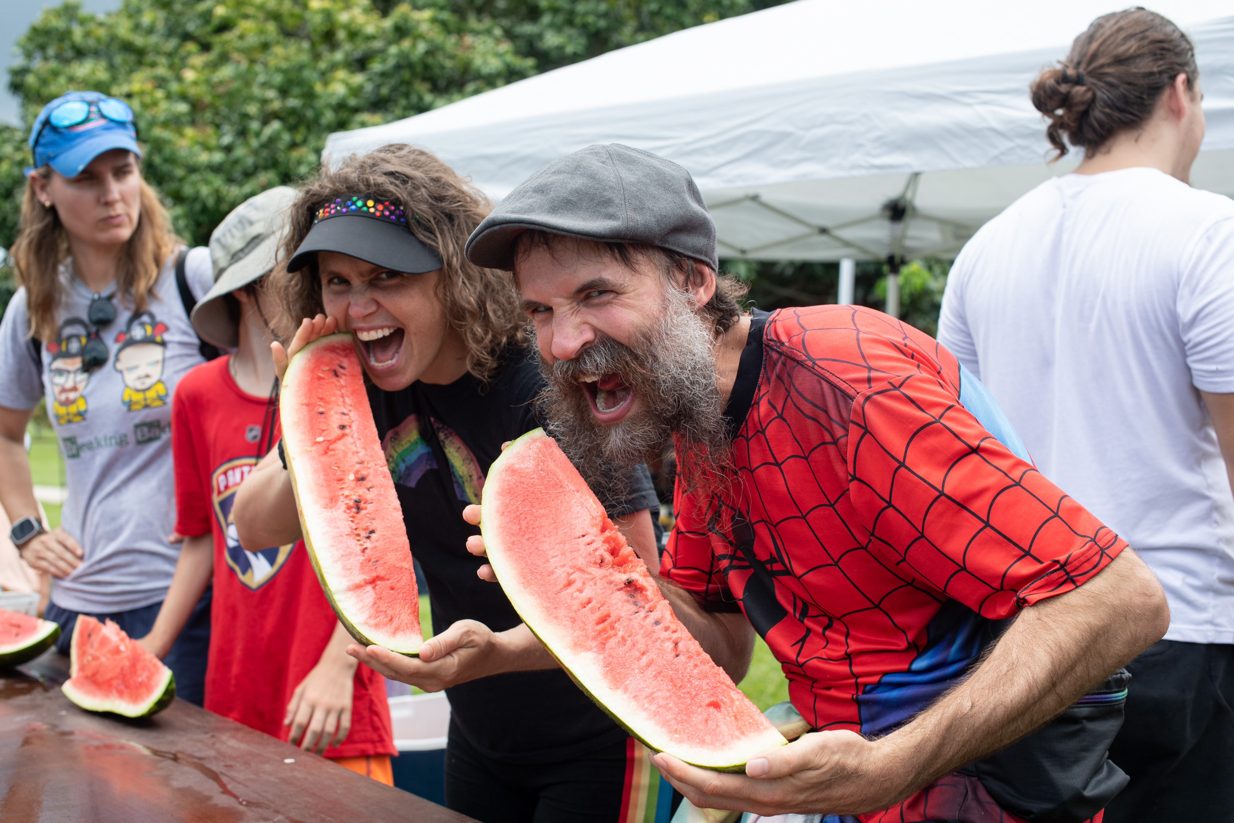Two people pretending to bite watermelon slices before a eating contest at an outdoor event.