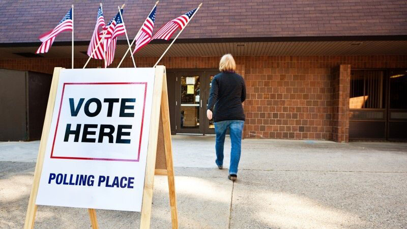 Sign in front of building reads "Vote Here" Polling Place. Male walking towards building