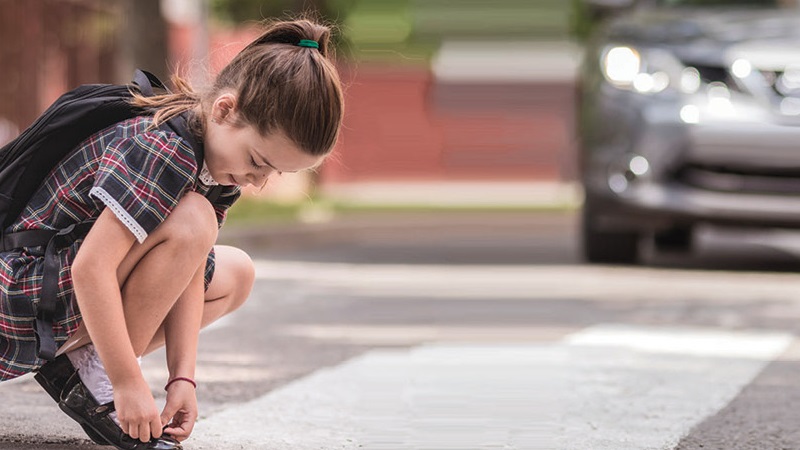 Image of a girl tying het shoe at a cross walk while a car is approaching.